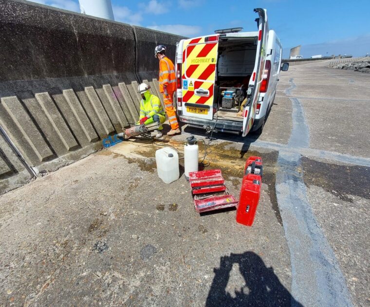Two highway maintenance workers in high-visibility clothing carrying out repairs beside a service van on a concrete coastal promenade.