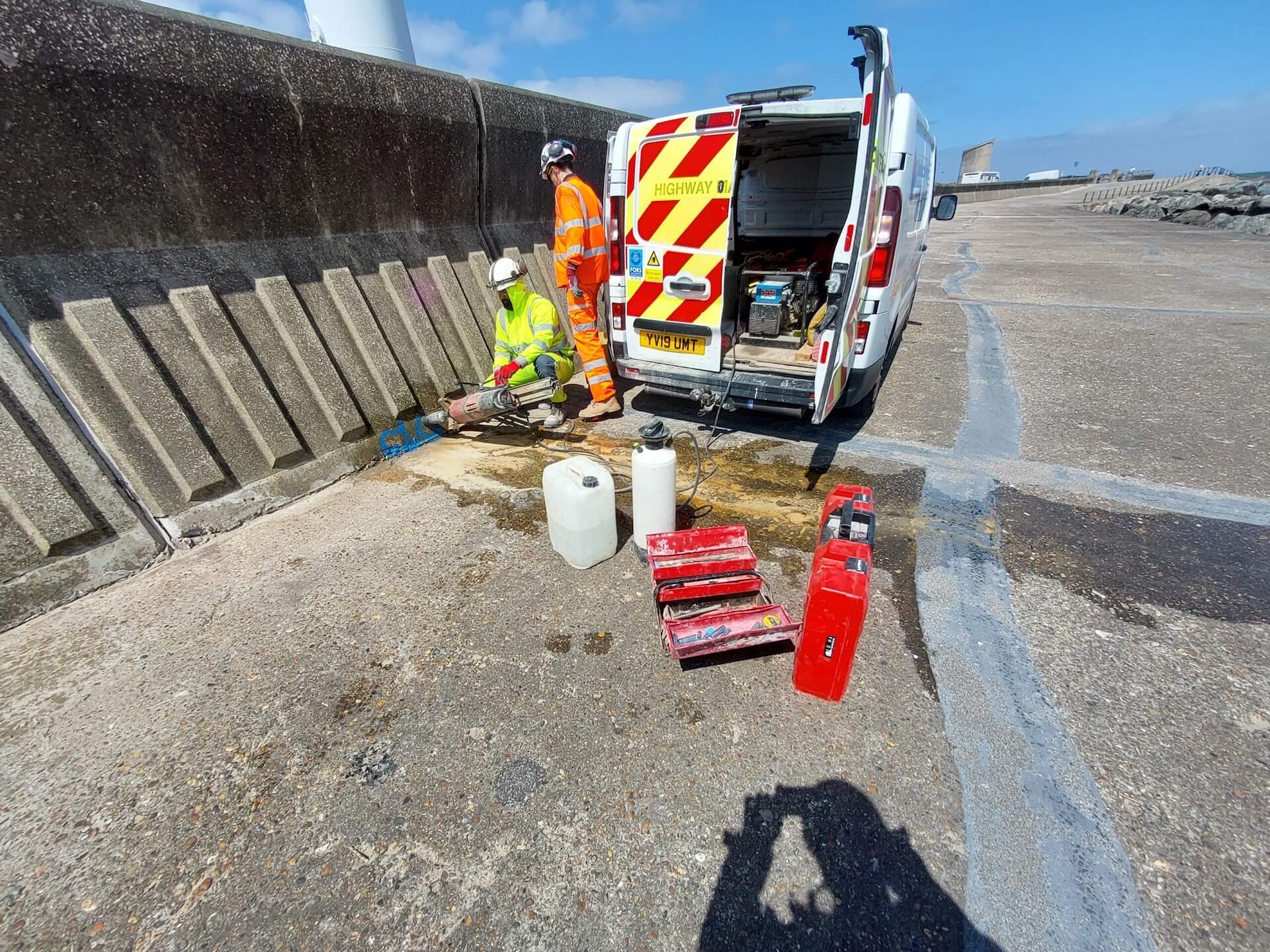Two highway maintenance workers in high-visibility clothing carrying out repairs beside a service van on a concrete coastal promenade.